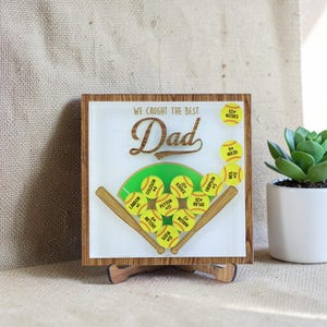 May include: A wooden sign with a softball theme, featuring the words "WE CAUGHT THE BEST Dad". The design includes a softball field with yellow softballs and names. The sign is displayed on a wooden stand, with a wooden frame.