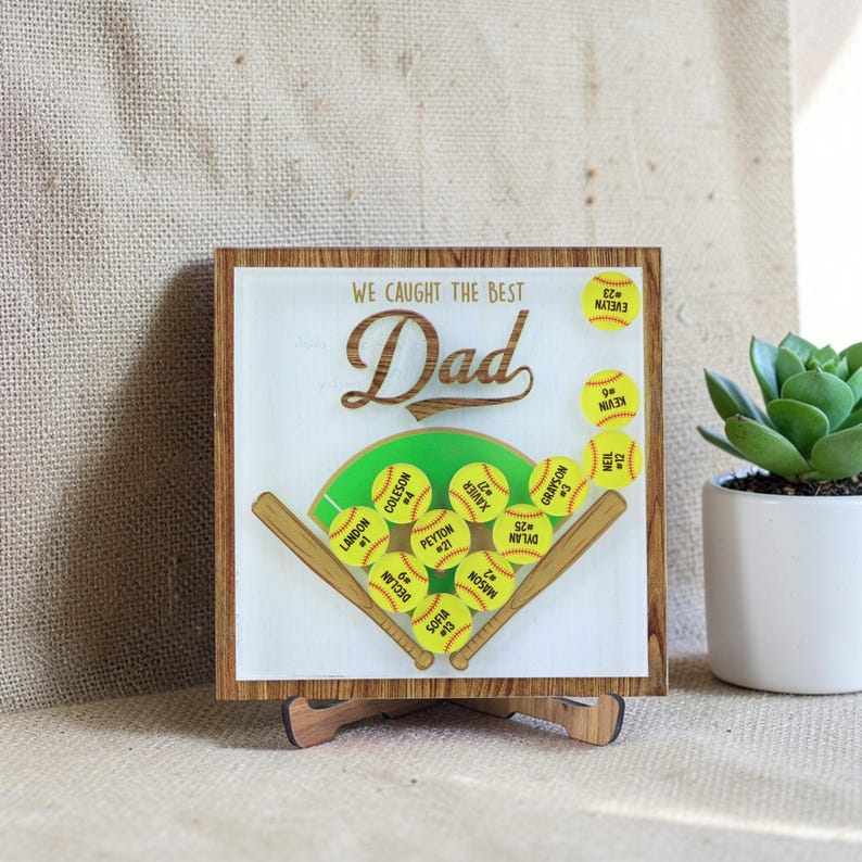 May include: A wooden sign with a softball theme, featuring the words "WE CAUGHT THE BEST Dad". The design includes a softball field with yellow softballs and names. The sign is displayed on a wooden stand, with a wooden frame.