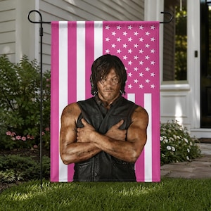 May include: A garden flag featuring a pink and white American flag design with a portrait of a man in a black vest with his arms crossed. The flag is displayed on a black metal stand, set against a backdrop of a house and green grass.