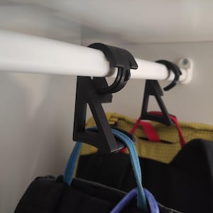 May include: Black plastic closet rod hangers are attached to a white closet rod. The hangers are holding backpacks with blue, red, and yellow accents. The closet rod is mounted inside a white closet.