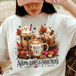 Könnte beinhalten: Ein cremefarbenes T-Shirt mit einer Grafik aus drei Kaffeetassen, Geistern, Kürbissen und Herbstblättern. Der Text lautet "Sippin' Magic", "Hello, Fall!" und "Autumn Leaves & Coffee Please". Das Design enthält Zimtstangen und kleine Sterne.