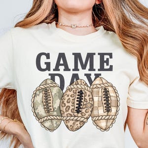 Könnte beinhalten: Ein cremefarbenes T-Shirt mit dem Schriftzug "GAME DAY" in dunkelgrauen Blockbuchstaben über drei Illustrationen von Footballs. Die Footballs zeigen verschiedene Muster: eines mit grünem Camouflage, eines mit Leopardenmuster und eines mit beigem Karomuster.