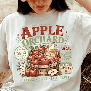 Könnte beinhalten: Ein cremefarbenes T-Shirt mit einem Grafikdruck eines Holzkörpers voller roter Äpfel und einer Glasflasche Apfelwein. Der Text lautet APPLE ORCHARD, FRESH DAILY PICKED, SUPPORT LOCAL, ORCHARD FAVORITE, U-PICK APPLES, APPLES CIDER FALL LEAVES.