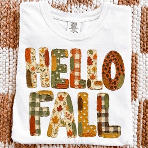 Könnte beinhalten: Ein weißes T-Shirt mit dem Text HELLO FALL in großen Blockbuchstaben. Jeder Buchstabe hat ein einzigartiges Muster, darunter Herbstblätter, orange-weißes Vichy-Karo, grüne Punkte, Leopardenmuster sowie grünes, oranges und braunes Karomuster.
