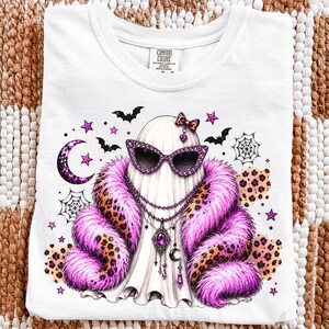 Könnte beinhalten: Ein weißes T-Shirt mit einer Grafik eines Geistes, der eine violette Sonnenbrille mit Leopardenmuster, eine rosa-leopardfarbene Kunstfellstola und eine mehrlagige Halskette mit einem violetten Edelstein trägt, umgeben von Fledermäusen und Sternen.