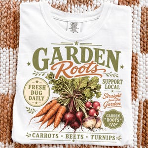 May include: A white t-shirt featuring a graphic print of carrots, beets, and turnips with the text "GARDEN Roots", "FRESH DUG DAILY", "SUPPORT LOCAL", "Straight from the Garden", "GARDEN ROOTS", and "CARROTS • BEETS • TURNIPS" in green and orange.