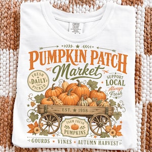 Könnte beinhalten: Ein weißes T-Shirt mit einem Grafikdruck eines hölzernen Wagens, der mit orangefarbenen Kürbissen und Zierkürbissen gefüllt ist. Der Text lautet PUMPKIN PATCH Market, FRESH DAILY PICKED, SUPPORT LOCAL Always Fresh, EST. 1958, FARM FRESH PUMPKINS, GOURDS VINES AUTUMN HARVEST.