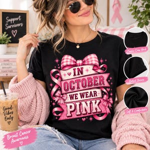 Op de afbeelding: Een zwart T-shirt met korte mouwen en een grafisch ontwerp met roze linten en de tekst In October We Wear Pink. Het shirt heeft een klassieke ronde hals en opgerolde mouwen. Het ontwerp bevat een grote roze gingham strik en kleine hartjes.