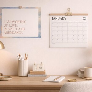 Può includere: Una scrivania per l'home office con una lampada color oro, un calendario e una stampa incorniciata con la scritta "I AM WORTHY OF LOVE, RESPECT AND ABUNDANCE". Una tazza bianca è appoggiata su un sottobicchiere accanto a un vaso con fiori secchi.