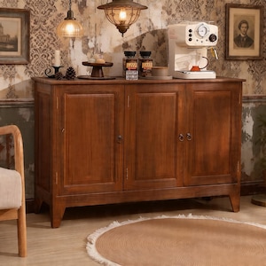 May include: A dark brown wooden cabinet with three doors, topped with a coffee machine, jars, and decorative items. The cabinet is set against a patterned wallpaper, with a round rug on the floor.