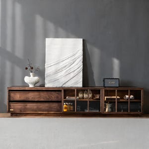 Puede incluir: Un mueble de entretenimiento de madera oscura con cajones, estantes abiertos y un armario. Una obra de arte blanca texturizada y un jarrón blanco con flores secas encima. Un pequeño altavoz y varios objetos decorativos se exhiben.