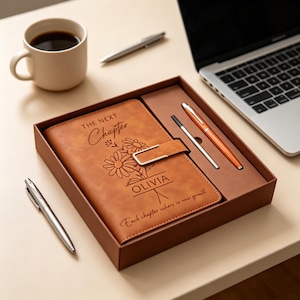 Può includere: Un set di diario in pelle marrone con una penna abbinata e una penna d'argento, presentato in una confezione regalo. Il diario è impresso con le parole "The Next Chapter" e "Olivia", insieme a disegni floreali. Una tazza di caffè e un laptop sono anche nell'immagine.