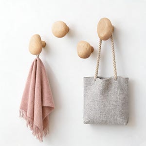 May include: Four wooden wall hooks with a natural finish are mounted on a white wall. A pink fringed towel hangs from one hook, and a gray tote bag with rope handles hangs from another. The hooks have a rounded, organic shape.