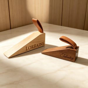 Könnte beinhalten: Zwei Türstopper aus Holz mit Ledergriffen. Einer ist hell mit der Gravur "Lorraine", der andere ist dunkler mit der Gravur "Talaskey". Beide sind keilförmig und liegen auf einer hellen Oberfläche.