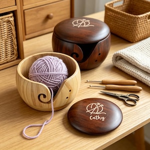 May include: Wooden yarn bowls and accessories. A light-coloured wooden yarn bowl holds a ball of lavender yarn. A dark brown wooden yarn bowl with a lid and a matching coaster are also present. Crochet hooks and scissors are nearby.