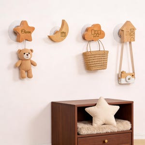 May include: Wooden wall hooks in star, moon, cloud, and house shapes. The star hook has the name "Owen" engraved. A teddy bear, a woven basket, and a toy camera hang from the hooks. A star-shaped pillow rests on a wooden cabinet.