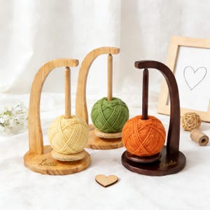 May include: Three wooden yarn holders, each with a different coloured ball of yarn: yellow, green, and orange. The holders have a curved arch design and a central dowel. The bases are round, with names "Lucas" and "Charlie" engraved on two.