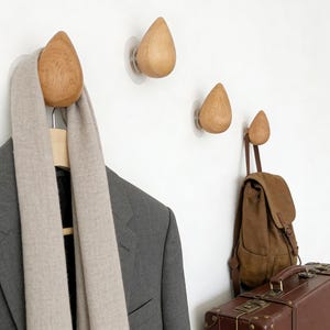 Peut inclure: Crochets muraux en forme de goutte d'eau en bois avec ventouses transparentes. Un crochet maintient un blazer gris et une écharpe beige sur un cintre en bois. Un sac à dos marron et une valise sont également visibles.