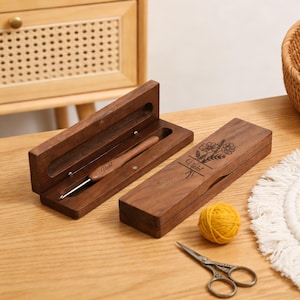 Puede incluir: Un juego de ganchillo y estuche de madera. El estuche marrón oscuro está grabado con diseños florales y el nombre "Violet". También hay una madeja de lana amarilla y unas tijeras pequeñas, lo que sugiere un tema artesanal.