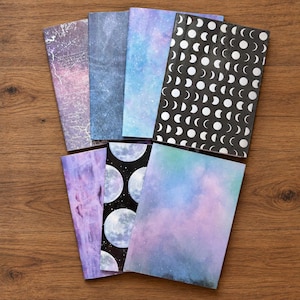 May include: A collection of seven rectangular notebooks featuring various celestial and space-themed covers. Designs include purple and blue nebula patterns, a black cover with white moon phases, and a black cover with large, detailed full moons.