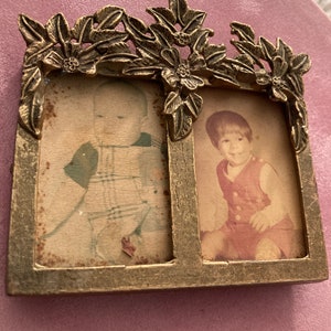 May include: A vintage double photo frame with a floral design. The frame is made of gold-colored metal and holds two small, rectangular photos. The frame is placed on a pink surface.