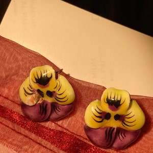 May include: A pair of purple and yellow flower-shaped earrings with black accents. The earrings are on a red and white fabric background.