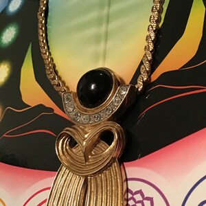 Puede incluir: Un collar de cadena dorada con un colgante que presenta una piedra negra rodeada de diamantes de imitaci&oacute;n transparentes. El colgante tiene la forma de una figura estilizada con un dise&ntilde;o fluido.