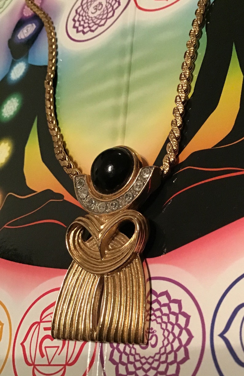 Puede incluir: Un collar de cadena dorada con un colgante que presenta una piedra negra rodeada de diamantes de imitaci&oacute;n transparentes. El colgante tiene la forma de una figura estilizada con un dise&ntilde;o fluido.