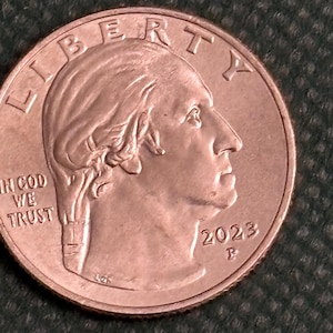 May include: A copper-colored United States quarter dollar coin with the words "LIBERTY" and "IN GOD WE TRUST" inscribed on the obverse. The coin features a profile of a woman's face and the year "2023" with the mint mark "P" below it.