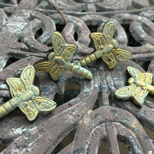 May include: Four antique-style dragonfly drawer pulls with a distressed gold and green finish. The dragonfly hardware is made of metal and is arranged on a weathered, brown metal surface.