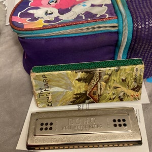 May include: A silver harmonica with the words "ECHO BELL METAL REEDS REGISTERED" engraved on the front. The harmonica is sitting on a white surface next to a book titled "The ECHO HARP" and a purple backpack with a My Little Pony design.