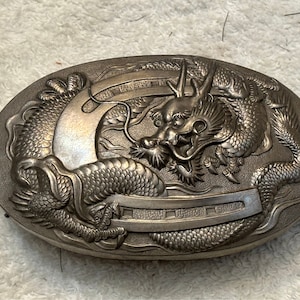 May include: Oval-shaped, silver-toned metal box with a detailed dragon relief. The dragon is centered, with its head and body curving around the box. The box has a textured surface and a decorative border.