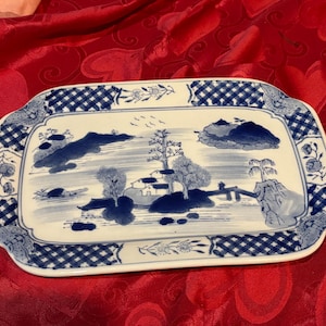 May include: Rectangular ceramic serving tray with a blue and white landscape design. The tray features a detailed illustration of a serene landscape with trees, houses, and water. The border has a blue checkered pattern.