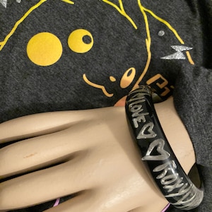 May include: Black bangle bracelet with silver text and heart designs. The bracelet is worn on a mannequin hand. The text on the bracelet reads "LOVE ROXY". The background is a gray shirt with a yellow Pikachu graphic.