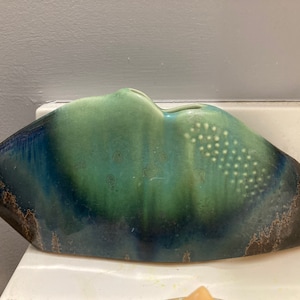 May include: A ceramic vase with a unique, abstract shape. The vase is glazed in shades of blue, green, and brown, with a speckled pattern on the surface.