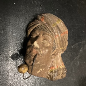 May include: A brown, vintage-style figurine of a man's head wearing a turban. The figurine has a pipe in its mouth and is painted with red and yellow accents. The overall design suggests a historical or cultural artifact.
