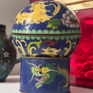 May include: A cylindrical cloisonné container featuring a vibrant blue base with a green dragon motif. The lid is a matching blue sphere decorated with yellow and pink floral patterns, finished with brass edges and intricate enamel detailing.