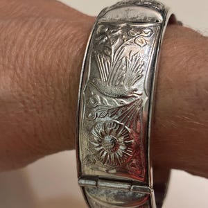 May include: A silver hinged bracelet with detailed floral and bird engravings. The bracelet is rectangular in shape and features a clasp closure. The design includes a bird, flowers, and foliage. The bracelet is approximately 1 inch wide.