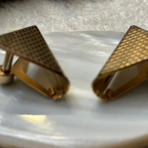 May include: A pair of gold-toned cufflinks with a geometric design. Each cufflink features a textured, triangular top and a curved, rectangular base. The cufflinks are displayed on a white, textured surface.