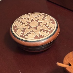 May include: A round, wooden trinket box with a starburst design on the lid. The lid features a light brown star pattern on a darker brown background. The box has a dark brown base with black and brown accents.