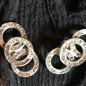 May include: Two pairs of silver-toned earrings. Each earring features a design of interlocking circles, encrusted with small, clear gemstones. The earrings are set against a dark, textured background, highlighting their sparkle.