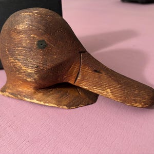 May include: A wooden duck head sculpture with a long beak and a dark brown finish. The sculpture has a small black dot for an eye and is set against a pink background. The duck head is likely a decorative item.