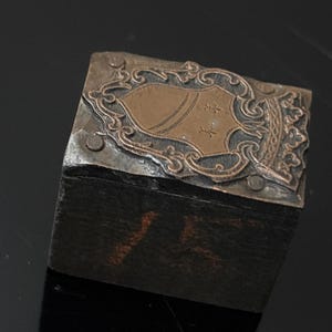 May include: A small, wooden box with a metal top. The top features a decorative design with a copper-colored shield and intricate scrollwork.