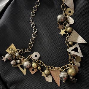 May include: A multi-strand necklace with a mix of gold, silver, and copper-toned metal charms. The charms include geometric shapes, stars, hearts, and spheres. The necklace has a chain closure and is displayed on a black surface.