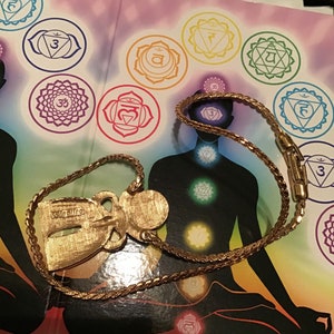 Puede incluir: Un collar de tono dorado con un colgante que presenta una figura de &aacute;ngel estilizada. El colgante tiene la palabra "valentino" inscrita en &eacute;l. El collar est&aacute; sobre un libro con una ilustraci&oacute;n colorida de una persona sentada en una postura meditativa con siete chakras.