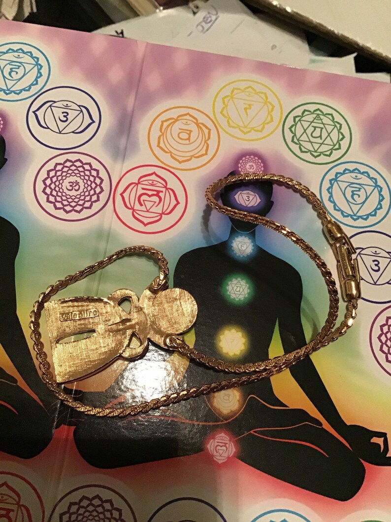 Puede incluir: Un collar de tono dorado con un colgante que presenta una figura de &aacute;ngel estilizada. El colgante tiene la palabra "valentino" inscrita en &eacute;l. El collar est&aacute; sobre un libro con una ilustraci&oacute;n colorida de una persona sentada en una postura meditativa con siete chakras.