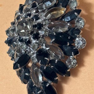 May include: A vintage brooch with a cluster design. The brooch features a variety of faceted stones in shades of black, gray, and clear, set in a silver-tone metal. The stones are arranged in a layered, asymmetrical pattern, creating a sparkling, eye-catching accessory.