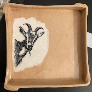 May include: Square ceramic tray with a tan and white goat illustration. The tray has a light tan color with a cracked texture. The goat is black and white and is on a white background.