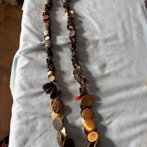 May include: A long necklace made of various buttons in different colors, shapes, and sizes. The buttons are strung together on a cord and include black, brown, gold, white, and red buttons.