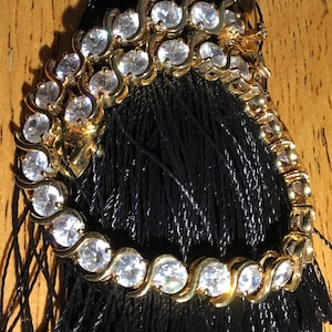 May include: A gold-toned bracelet with clear, round gemstones set in a wave pattern. The bracelet is displayed on a black tassel, creating a contrast of textures and colors. The clasp is visible, indicating a secure closure.